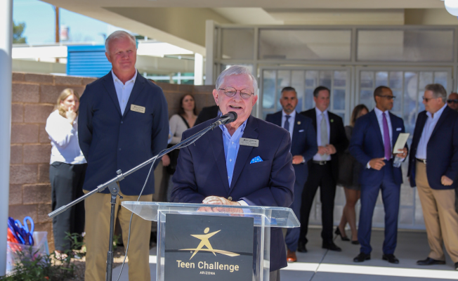 A man speaks at a podium labeled, while several people stand behind him at an outdoor event.