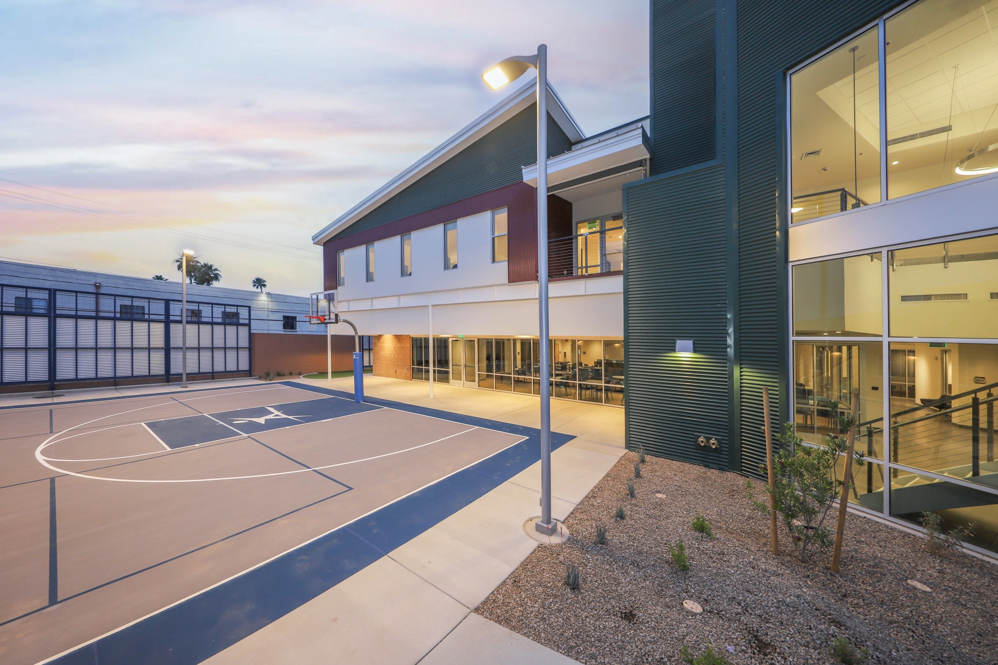 An outdoor basketball court with a light brown surface and three-point line, adjacent to a modern building featuring large windows and a green metal facade. The area is well-lit with street lights, surrounded by gravel and small shrubs.