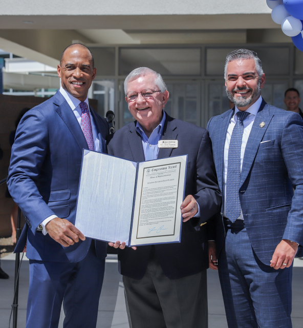 Three men are standing outdoors holding a large certificate. The man on the left is wearing a dark suit with a purple tie, the man in the center is in a black blazer, and the man on the right is in a checked suit. They are smiling at the camera.