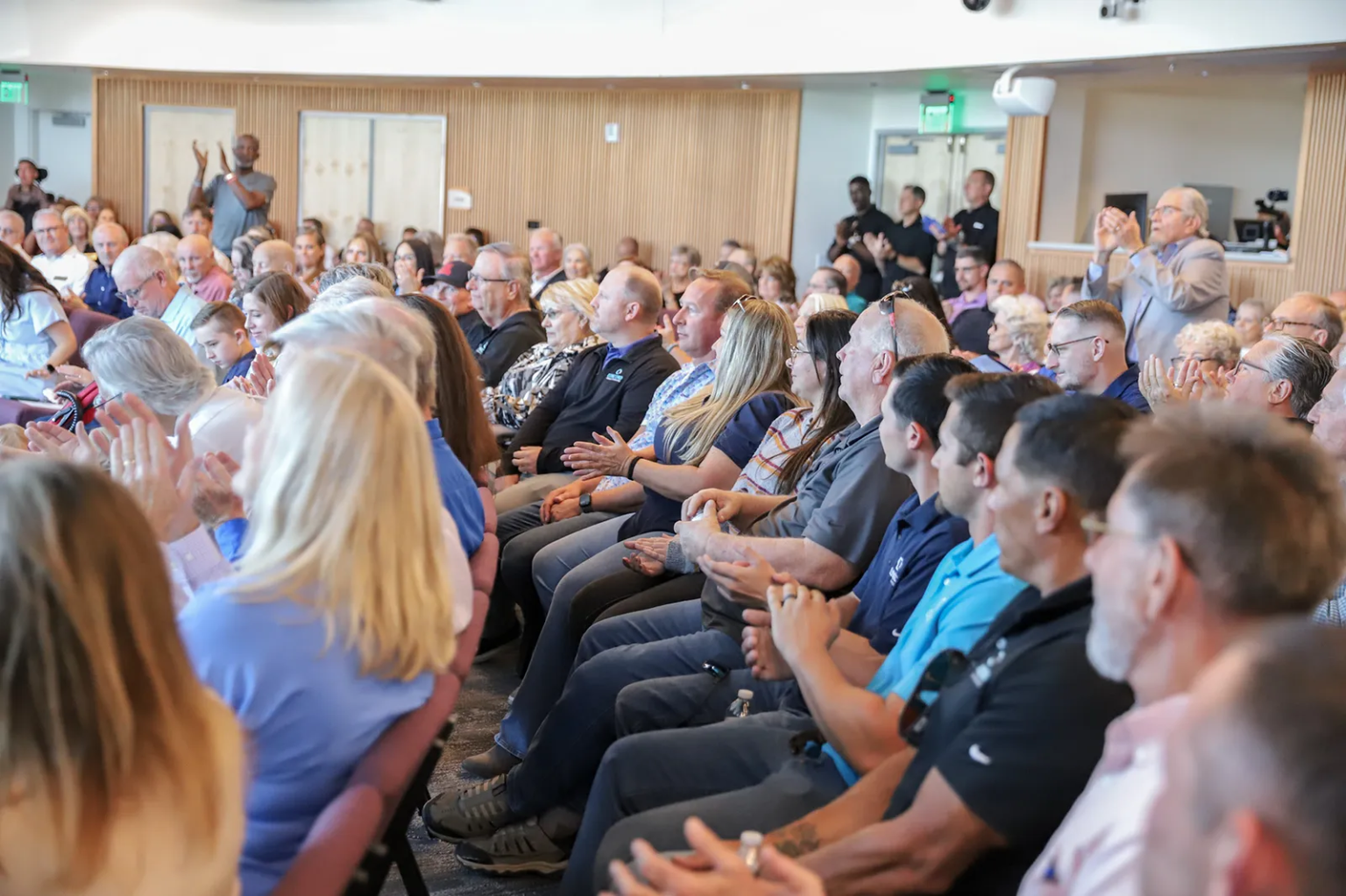 A diverse audience seated in a conference hall, clapping and engaging during an event, with a mix of ages and backgrounds visible.