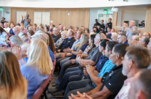 A diverse audience seated in a conference hall, clapping and engaging during an event, with a mix of ages and backgrounds visible.